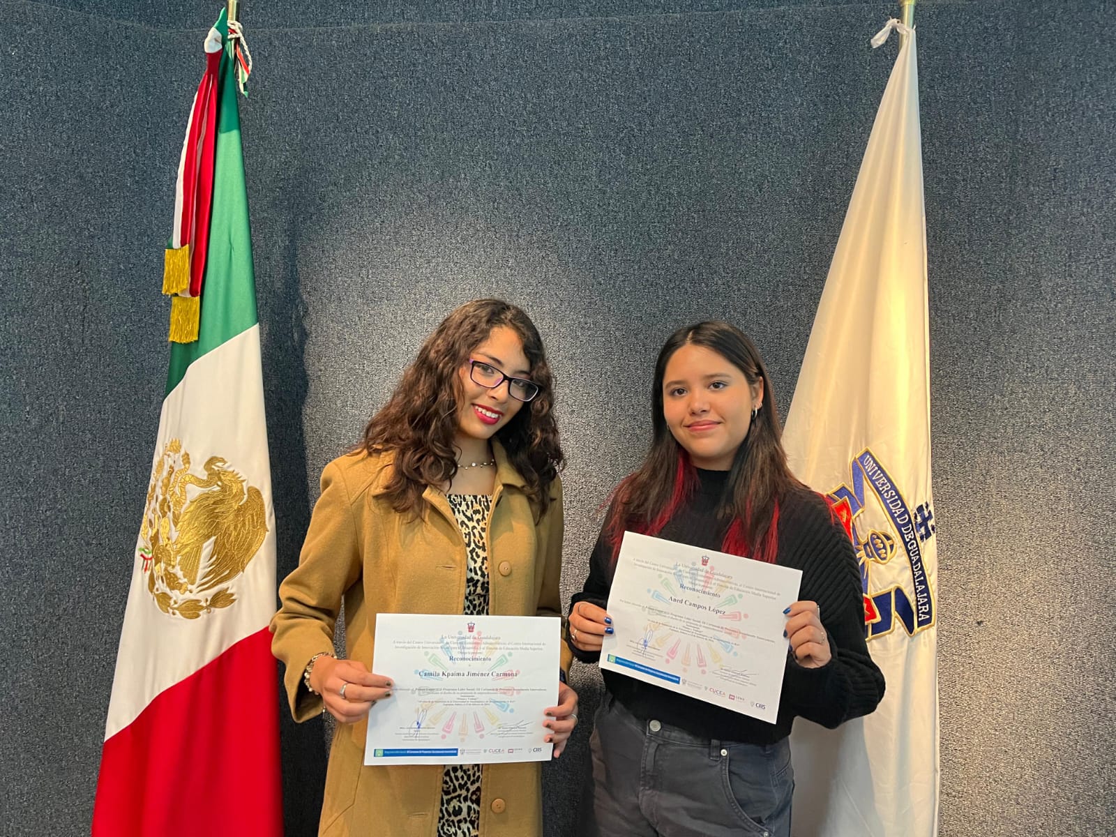 Dos mujeres de pie junto a bandera de México y U D G sostienen reconocimientos