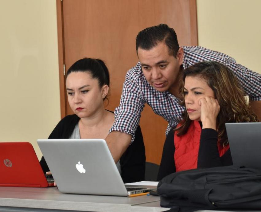 Dos mujeres sentadas frente a computadoras y hombre de pie les explica